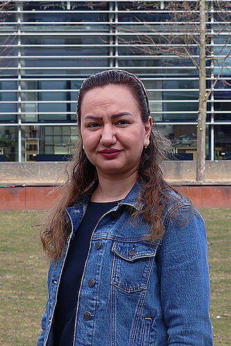 Porträt einer Frau im Freien vor einem modernen Hochschulgebäude. Sie trägt eine Jeansjacke und blickt in die Kamera.