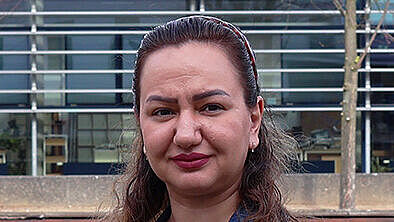 A portrait of a woman outdoors in front of a modern university building. She is wearing a denim jacket and looking at the camera.