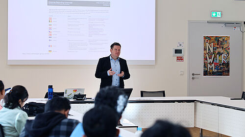 Ein Mann steht vor Studierenden vor einer Leinwand auf der eine Vorlesungsfolie zu sehen ist und hält eine Lehrveranstaltung ab.