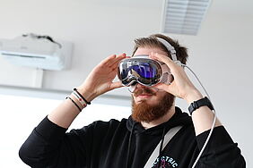 Ein junger Mann mit Vollbart setzt sich eine Apple Vision Pro Brille auf. Er trägt einen schwarzen Hoodie und Armbänder. Im Hintergrund ist ein heller Seminarraum mit Klimaanlage und Deckenbeleuchtung zu sehen.