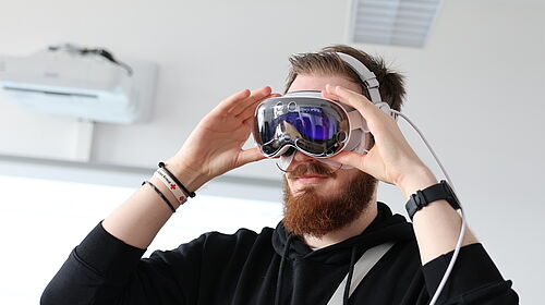 Ein junger Mann mit Vollbart setzt sich eine Apple Vision Pro Brille auf. Er trägt einen schwarzen Hoodie und Armbänder. Im Hintergrund ist ein heller Seminarraum mit Klimaanlage und Deckenbeleuchtung zu sehen.