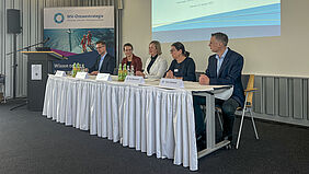 Die fünf Personen sitzen nebeneinander an einem Tisch. An der Wnad dahinter ist eine Präsentation mit dem Titel der Veranstaltung zu sehen.