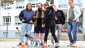 Studierende beobachten und steuern einen vierbeinigen Roboter auf dem Campus.