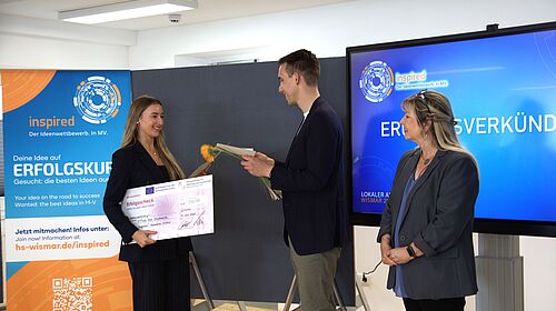 Die Masterstudentin Svea Bublys nimmt bei der Preisverleihung des Inspired-Wettbewerbs 2025 einen symbolischen Scheck und eine Blume entgegen. Neben ihr stehen ein Moderator sowie eine weitere Person. Im Hintergrund sind ein Roll-Up und ein Bildschirm mit der Aufschrift „Ergebnisverkündung“ zu sehen.