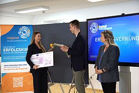 Die Masterstudentin Svea Bublys nimmt bei der Preisverleihung des Inspired-Wettbewerbs 2025 einen symbolischen Scheck und eine Blume entgegen. Neben ihr stehen ein Moderator sowie eine weitere Person. Im Hintergrund sind ein Roll-Up und ein Bildschirm mit der Aufschrift „Ergebnisverkündung“ zu sehen.