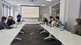 AIKnowIT final conference in Klaipėda Several project participants are seated at a U-shaped table in a modern seminar room. A speaker stands in front of a screen showing a presentation on the Interreg project AIKnowIT. The participants follow the presentation attentively, some working on their laptops.