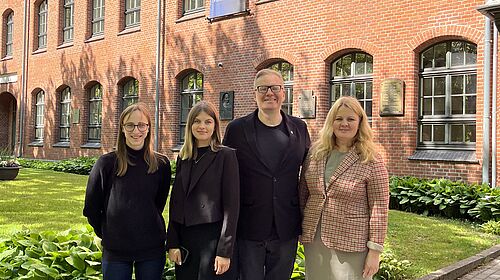 Vier Menschen stehen im freien vor einer grünen Wiese und einem rotgeklinkten Gebäude. Drei Frauen und ein Mann.