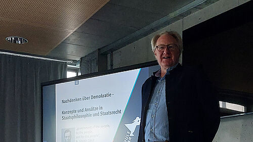 Die Person steht vor einem großen Bildschirm. Es ist der Oberkörper zu sehen und links davon eine digitale Präsentation, auf der das Logo der Hochschule Wismar zu sehen ist sowie der Titel „Nachdenken über Demokratie“.