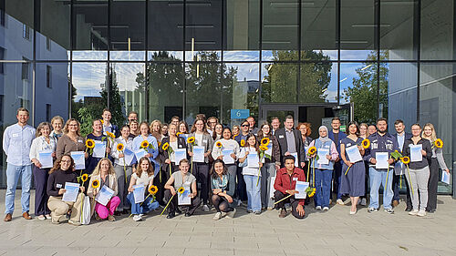 A group of people are standing or crouching in several rows in front of a modern glass building. Many are holding certificates and sunflowers in their hands.