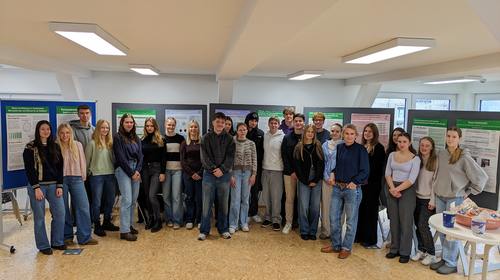 Studierendengruppe der Hochschule Wismar posiert vor einer Reihe von Präsentationstafeln mit wissenschaftlichen Postern im Ausstellungsraum. Die jungen Menschen stehen nebeneinander und blicken in die Kamera. Rechts im Vordergrund ein Tisch mit Gebäck und Kaffeetassen.