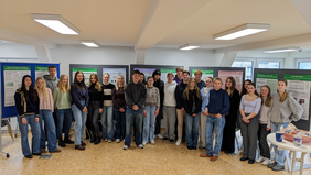 Studierendengruppe der Hochschule Wismar posiert vor einer Reihe von Präsentationstafeln mit wissenschaftlichen Postern im Ausstellungsraum. Die jungen Menschen stehen nebeneinander und blicken in die Kamera. Rechts im Vordergrund ein Tisch mit Gebäck und Kaffeetassen.