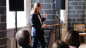 A woman in a dark blue blazer is giving a speech at a lectern in a hall with dark stone walls and windows. The audience is seated in front of her, their backs partially visible. She is gesturing with one hand to emphasise her words.