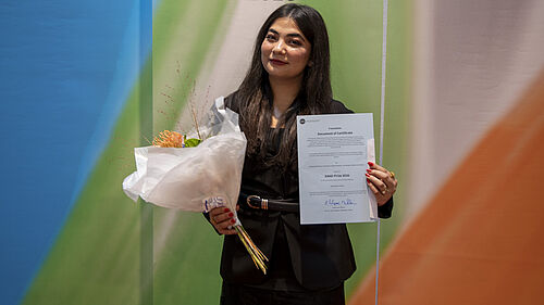 Frau in schwarzem Kleid hält Blumenstrauß und Urkunde vor einem Banner mit Schrift 'Feierliche Immatrikulation 2026'