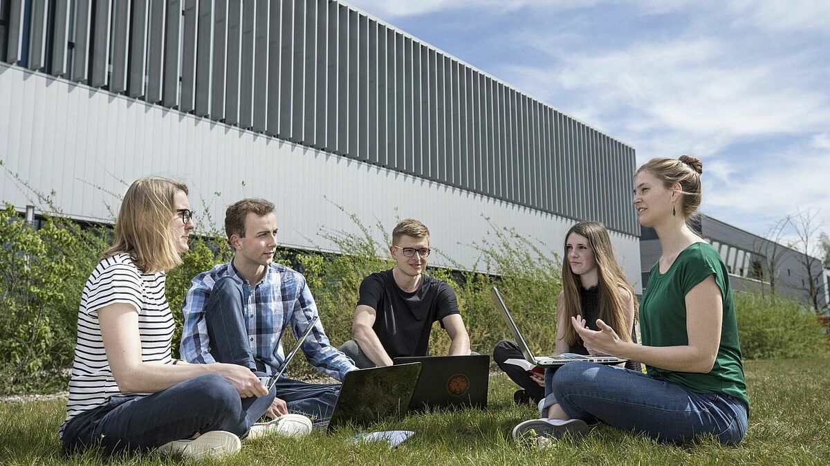 Es sind fünf Studierende mit Laptops zu sehen, die auf dem Rasen vor dem Haus 6 sitzen und miteinander sprechen. 