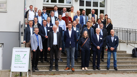 Group photo of around 30 participants at the 106th Federal Dean Conference on Economics at Wismar University of Applied Sciences. The people are standing on an outdoor staircase in front of a bright building with large windows. In the foreground on the left is a sign that reads ‘Welcome to the 106th Federal Dean Conference – Transfer and Change.’