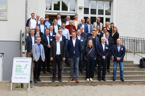 Group photo of around 30 participants at the 106th Federal Dean Conference on Economics at Wismar University of Applied Sciences. The people are standing on an outdoor staircase in front of a bright building with large windows. In the foreground on the left is a sign that reads ‘Welcome to the 106th Federal Dean Conference – Transfer and Change.’