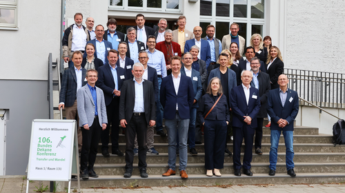 Group photo of around 30 participants at the 106th Federal Dean Conference on Economics at Wismar University of Applied Sciences. The people are standing on an outdoor staircase in front of a bright building with large windows. In the foreground on the left is a sign that reads ‘Welcome to the 106th Federal Dean Conference – Transfer and Change.’