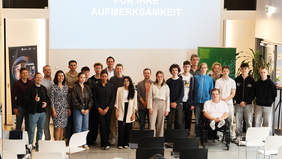Gruppenfoto mit rund 25 Personen, darunter Schülerinnen und Schüler, Jury und Hochschulvertretende. Sie stehen gemeinsam im Veranstaltungsraum des InnovationPort Wismar vor einer Leinwand mit der Aufschrift „[...] für Ihre Aufmerksamkeit“.