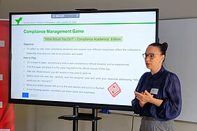 Eine Dozentin erklärt das „Compliance Management Game“ vor einem großen Bildschirm, der eine Spielanleitung mit Reflexionsfragen und Aufgaben zeigt.