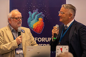 A: Die beiden mit Sakkos bekleideten Männer haben Mikrofone in der Hand und schauen sich an. Im Hintergrund befindet sich ein Banner mit einer Herzgrafik.