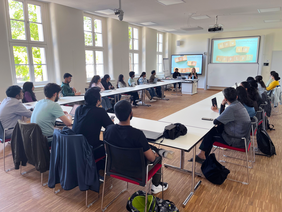 Students sit in a bright seminar room with large windows arranged around rectangular tables in a U-shape. Two female presenters stand at the front beside a table, presenting a coaching workshop titled “How to apply?” on two large screens. The participants listen attentively and take notes.
