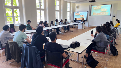 Students sit in a bright seminar room with large windows arranged around rectangular tables in a U-shape. Two female presenters stand at the front beside a table, presenting a coaching workshop titled “How to apply?” on two large screens. The participants listen attentively and take notes.