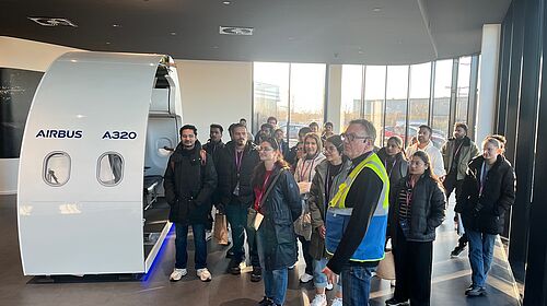 A group of students stands in front of a training module on aircraft production during a factory tour at Airbus.