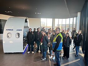 A group of students stands in front of a training module on aircraft production during a factory tour at Airbus.