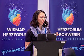 Die Professorin steht hinter einem Pult vor zwei farbigen Bannern, auf denen eine Herzgrafik zu sehen ist. Sie blickt in Richtung Publikum.